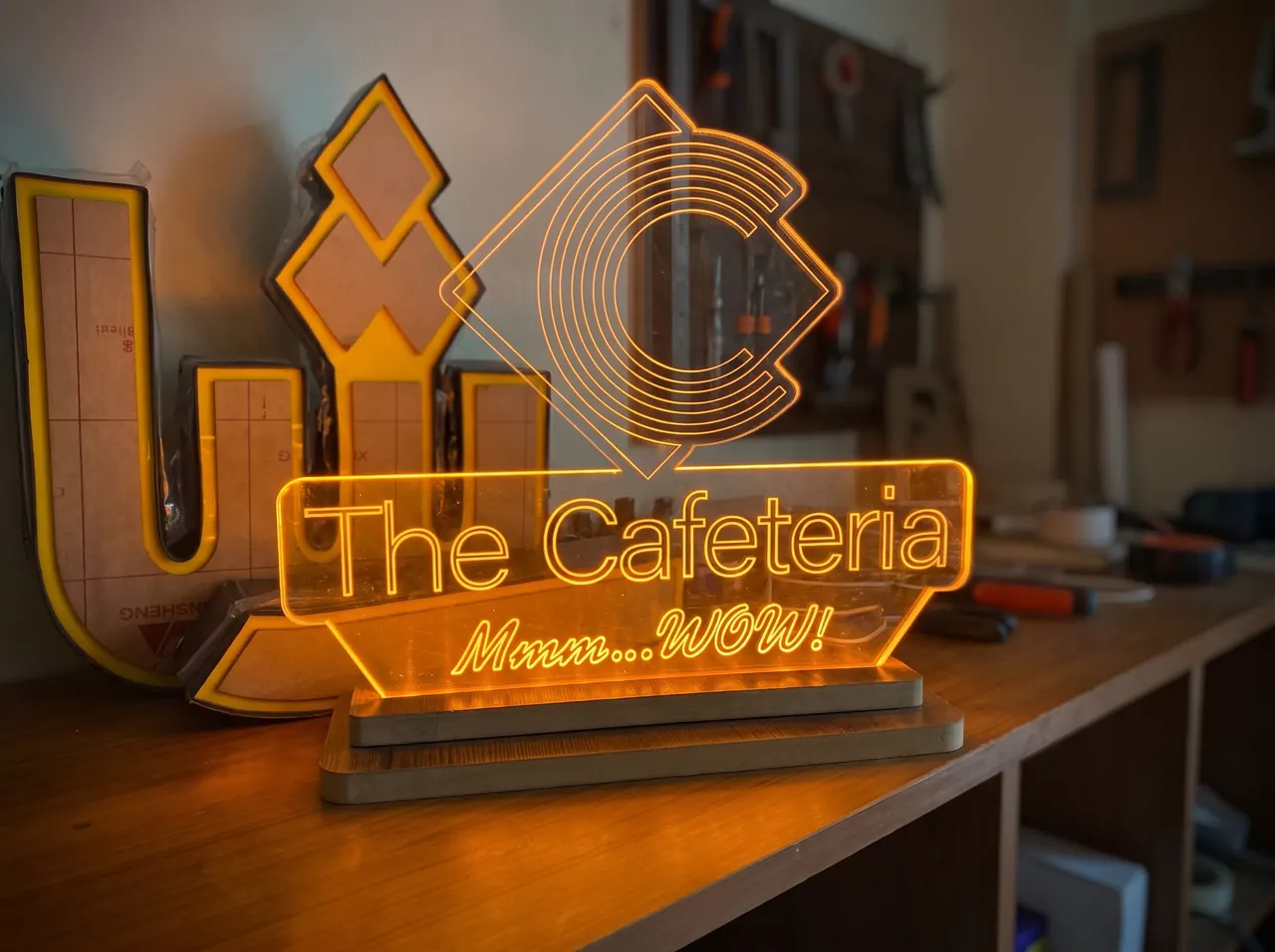Edge-lit amber acrylic counter sign reading The Cafeteria, Mmm WOW, glowing on a timber base in the workshop.